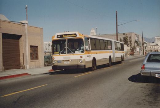 A white, yellow, and beige articulated bus with the destination 'Beverly Hills' is driving down a city street on a clear day, with a blue car parked nearby.