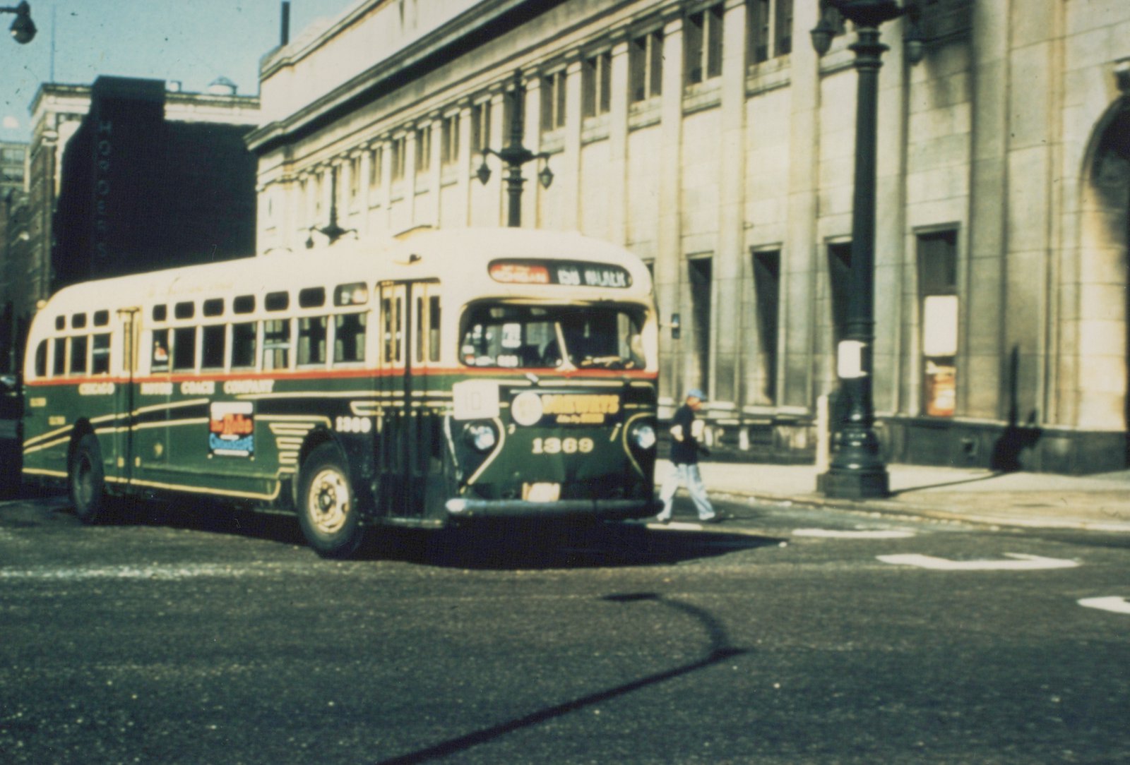 A green and cream Chicago Motor Coach Company bus, number 1369, is parked on a city street with a pedestrian walking nearby and a large stone building in the background.