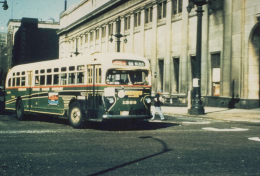 A green and cream Chicago Motor Coach Company bus, number 1369, is parked on a city street with a pedestrian walking nearby and a large stone building in the background.