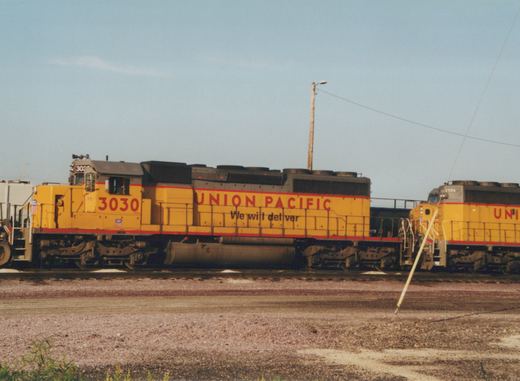 Side view of a yellow Union Pacific diesel locomotive, number 3030, sitting on tracks in a railroad yard under a clear blue sky.