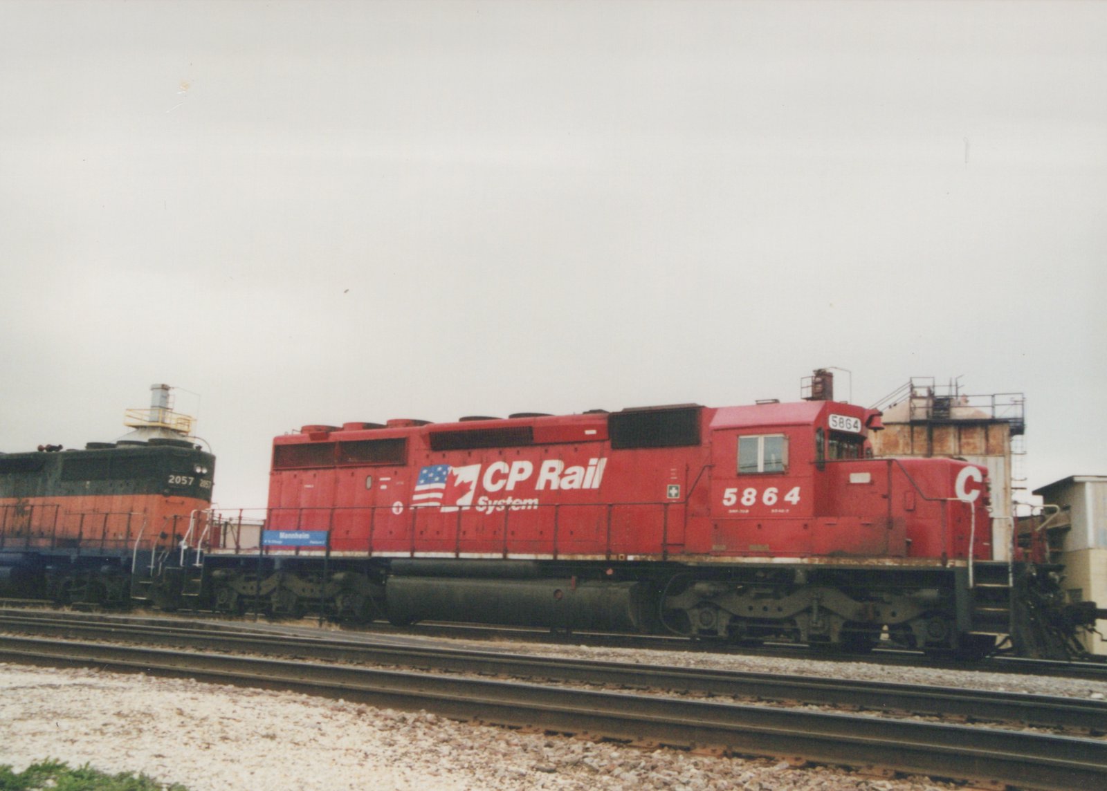 A red CP Rail locomotive numbered 5864 is parked on a track next to a dark green and orange Chicago and North Western locomotive numbered 2057, both at a yard with a blue sign reading 'Mannheim' in the background.