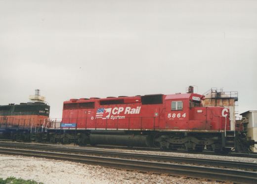A red CP Rail locomotive numbered 5864 is parked on a track next to a dark green and orange Chicago and North Western locomotive numbered 2057, both at a yard with a blue sign reading 'Mannheim' in the background.
