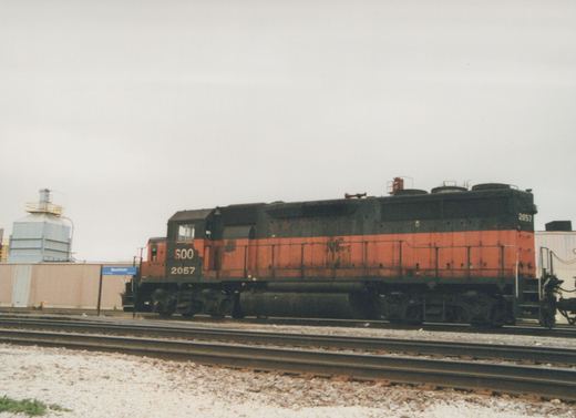 A side view of a SOO Line diesel locomotive, numbered 2057, parked on railroad tracks with a blue Mannheim station sign visible in the background.