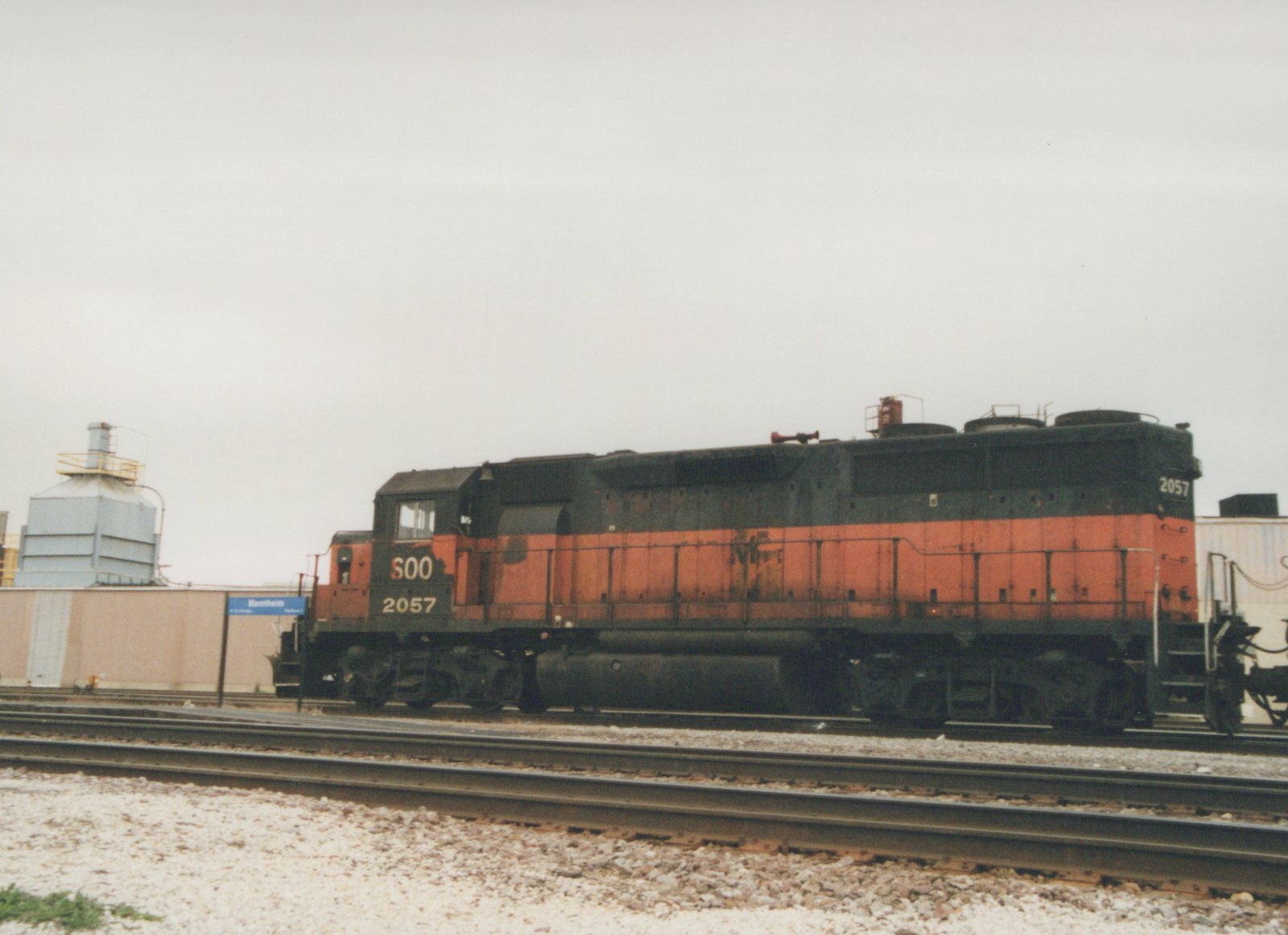 A side view of a SOO Line diesel locomotive, numbered 2057, parked on railroad tracks with a blue Mannheim station sign visible in the background.