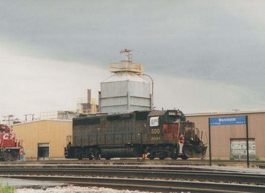 A black Soo Line locomotive, number 2041, is stationary on tracks at the Mannheim station, with a blue station sign and a red CP locomotive in the background.