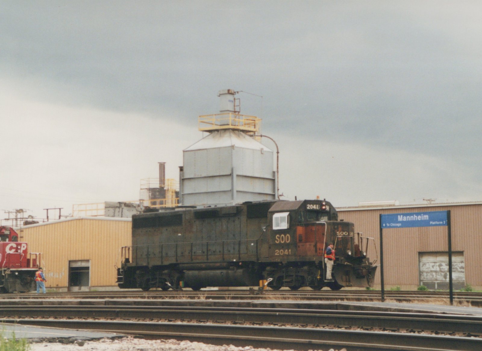 A black Soo Line locomotive, number 2041, is stationary on tracks at the Mannheim station, with a blue station sign and a red CP locomotive in the background.