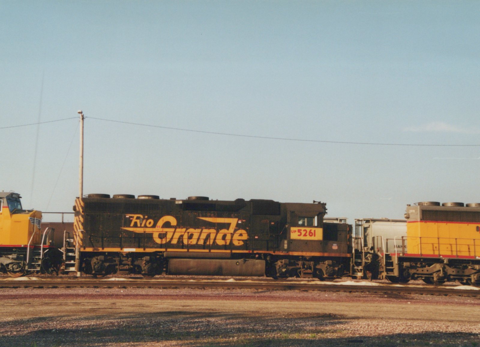 A side view of a black Union Pacific diesel locomotive with 'Rio Grande' written in yellow, numbered UP 5261, sitting on tracks in a gravel yard with another yellow locomotive nearby under a clear blue sky.