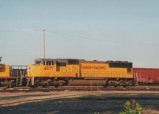 A yellow and red Union Pacific locomotive numbered 4077 is stationary on railroad tracks in a yard, with a clear blue sky above.
