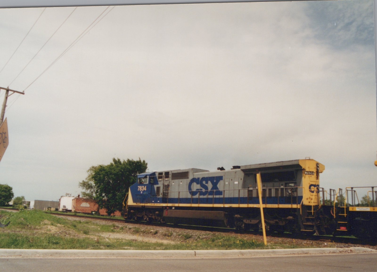 A CSX diesel locomotive numbered 7834 stands on the tracks beside a road, its gray, blue, and yellow livery visible under a cloudy sky.