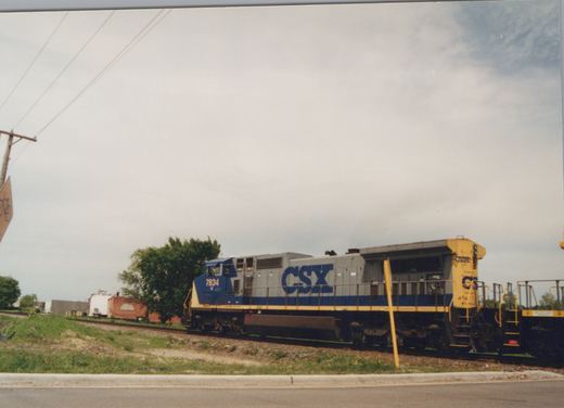 A CSX diesel locomotive numbered 7834 stands on the tracks beside a road, its gray, blue, and yellow livery visible under a cloudy sky.