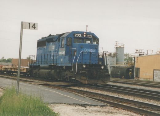 A blue diesel locomotive with Conrail and Railink lettering, numbered 203, sits on tracks in a railroad yard with industrial buildings and a trackside sign showing the number 14.