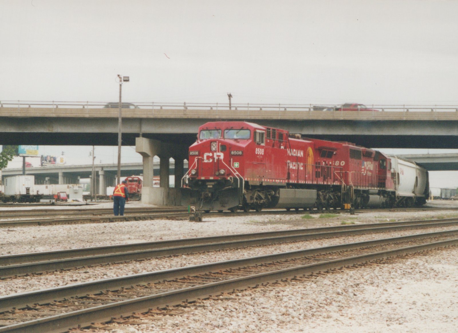 A red Canadian Pacific diesel locomotive numbered 8508 is on a set of tracks with a freight car behind it, a worker nearby, and a concrete overpass overhead.