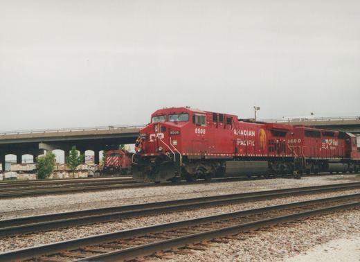A red Canadian Pacific locomotive with the number 8508 is shown on a railroad track, with another locomotive numbered 5680 behind it and a third one numbered 5724 in the background, all under a concrete bridge on an overcast day.