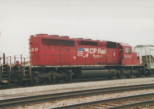 A red diesel locomotive with CP Rail System lettering and a dual flag logo is parked on tracks in a yard, with the number 5680 visible on the cab, and a white freight car in the background.