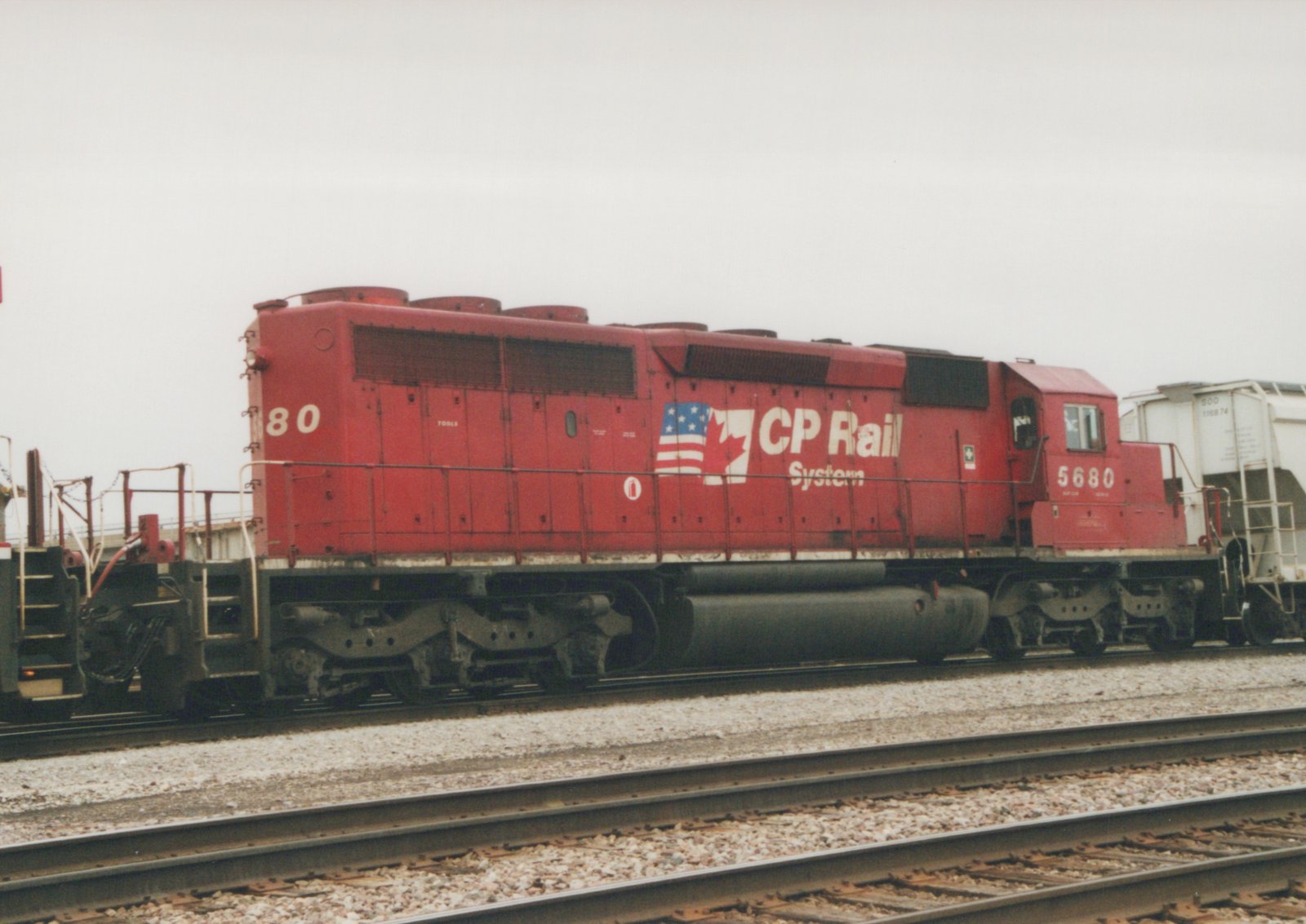 A red diesel locomotive with CP Rail System lettering and a dual flag logo is parked on tracks in a yard, with the number 5680 visible on the cab, and a white freight car in the background.