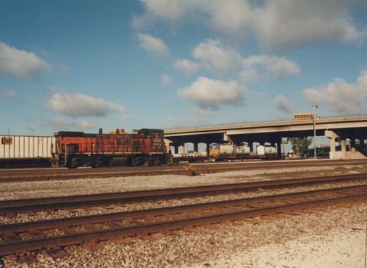 A SOO Line GP9 diesel locomotive, numbered 1553, is parked on a mainline track next to a white boxcar. In the background, a concrete overpass carries a yellow school bus over the tracks under a blue sky with white clouds.