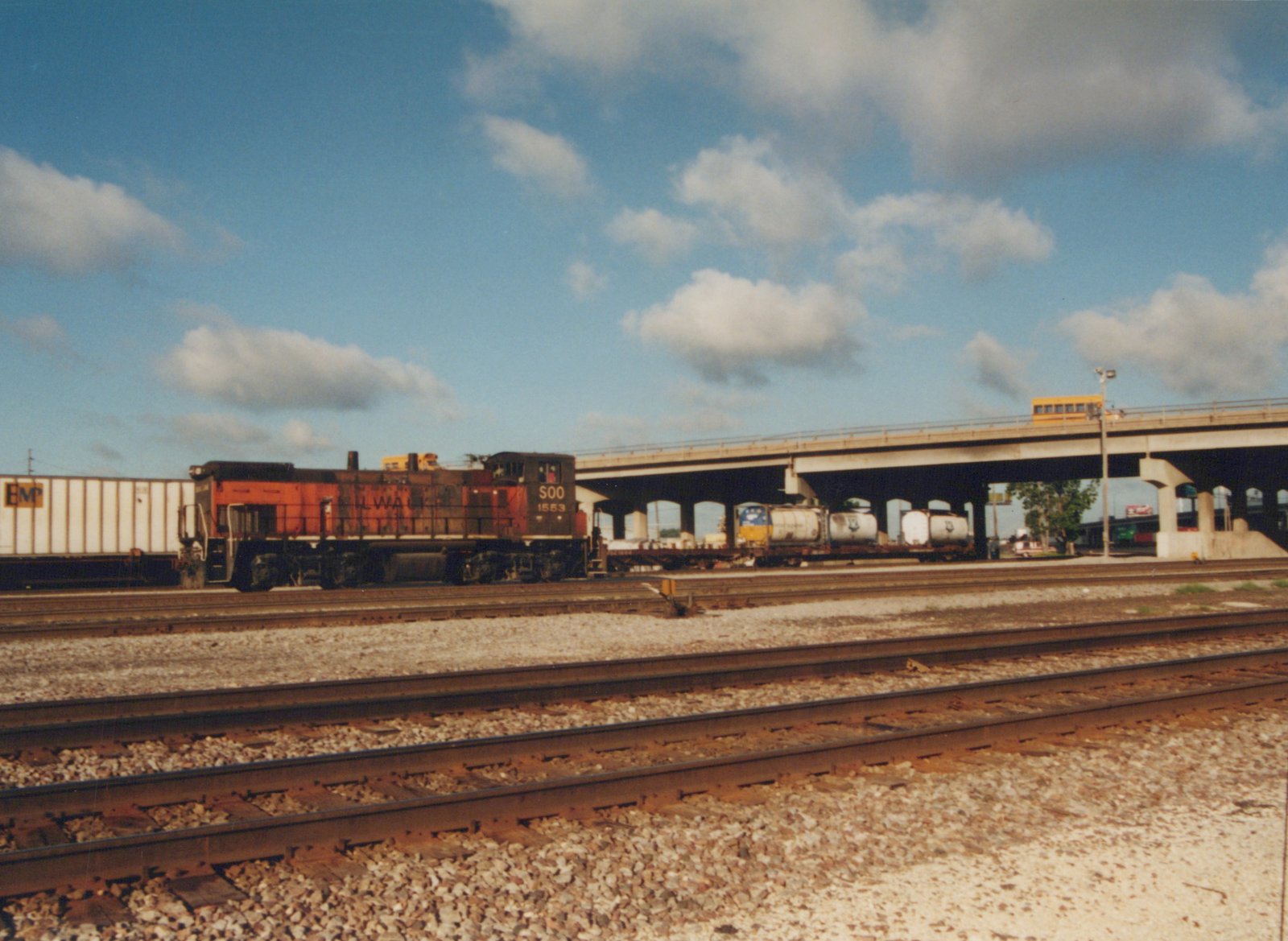 A SOO Line GP9 diesel locomotive, numbered 1553, is parked on a mainline track next to a white boxcar. In the background, a concrete overpass carries a yellow school bus over the tracks under a blue sky with white clouds.