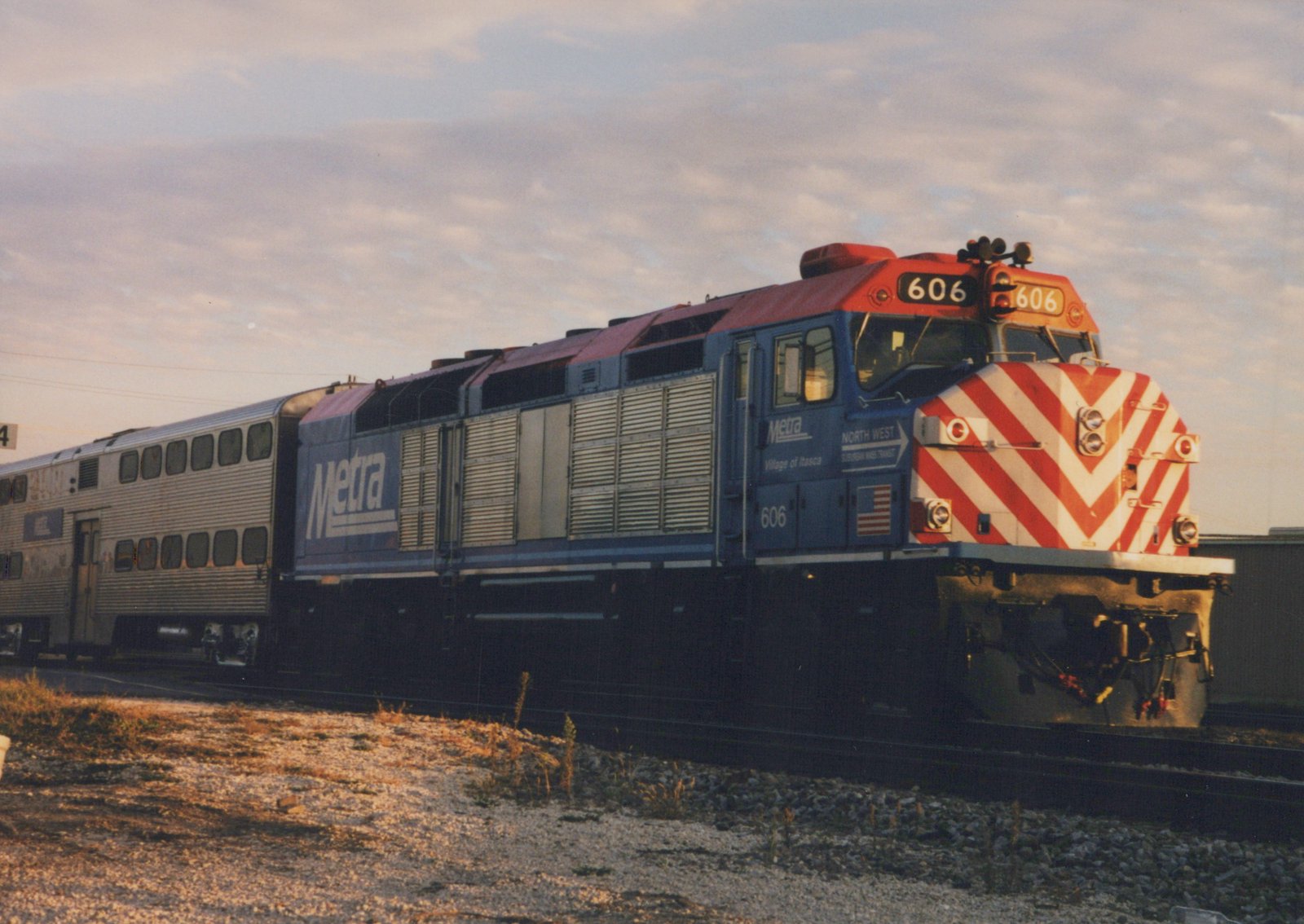 A blue and red Metra locomotive numbered 606 is pulling a train of silver commuter cars along a railroad track at dusk, with a cloudy sky above.