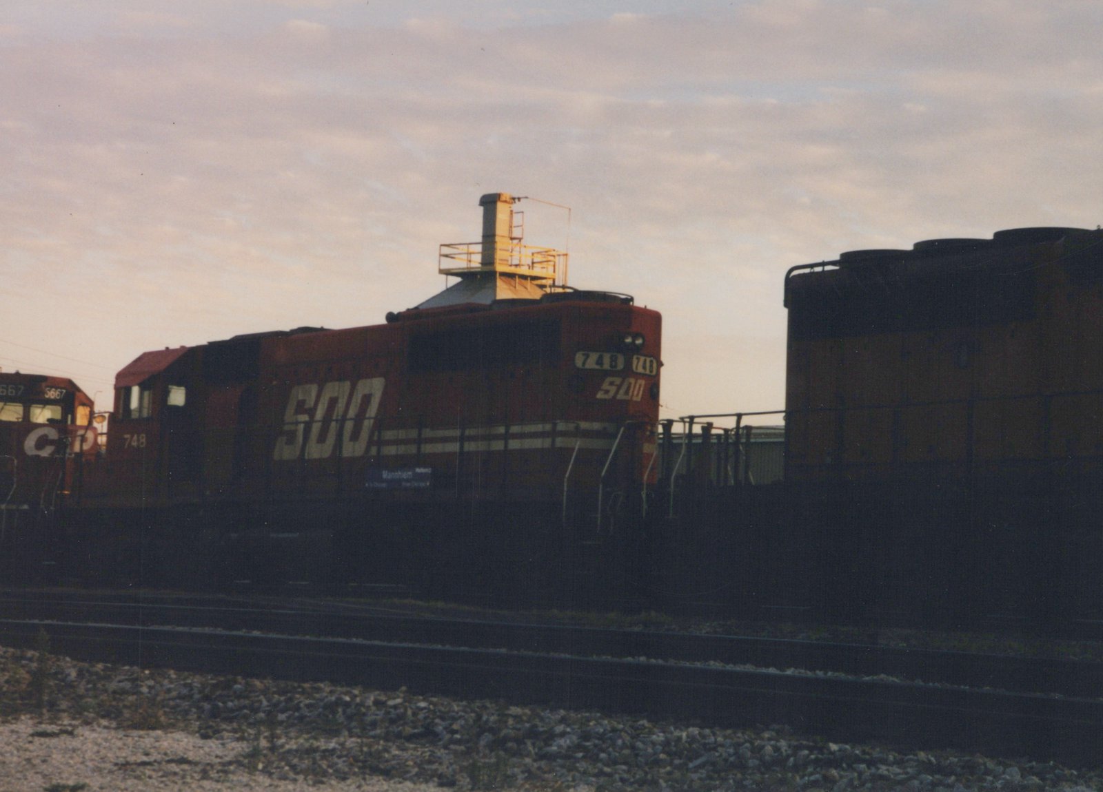 A maroon C&NW GP9 diesel locomotive, number 748, is parked in a rail yard at dusk, with another C&NW locomotive, number 5667, partially visible behind it.