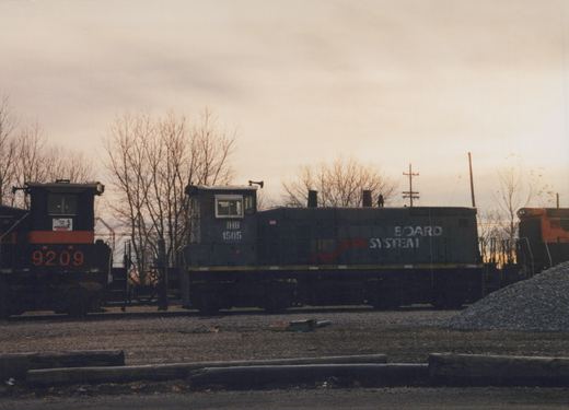 A blue Indiana Harbor Belt diesel locomotive with the number 1505 and the words BOARD SYSTEM on its side, parked in a gravel yard with bare trees and other locomotives nearby.