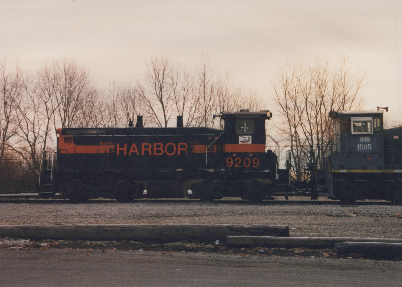 Side view of two diesel locomotives, one black with orange lettering and the other blue gray, parked on parallel tracks in a leafless winter landscape.