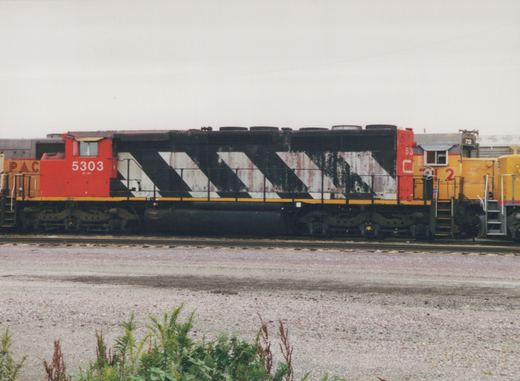 Side view of two diesel locomotives in a gravel yard: a red and black C&NW GP30r with number 5303, and a yellow Pacific Electric SW1200 with number 712, under a gray sky.