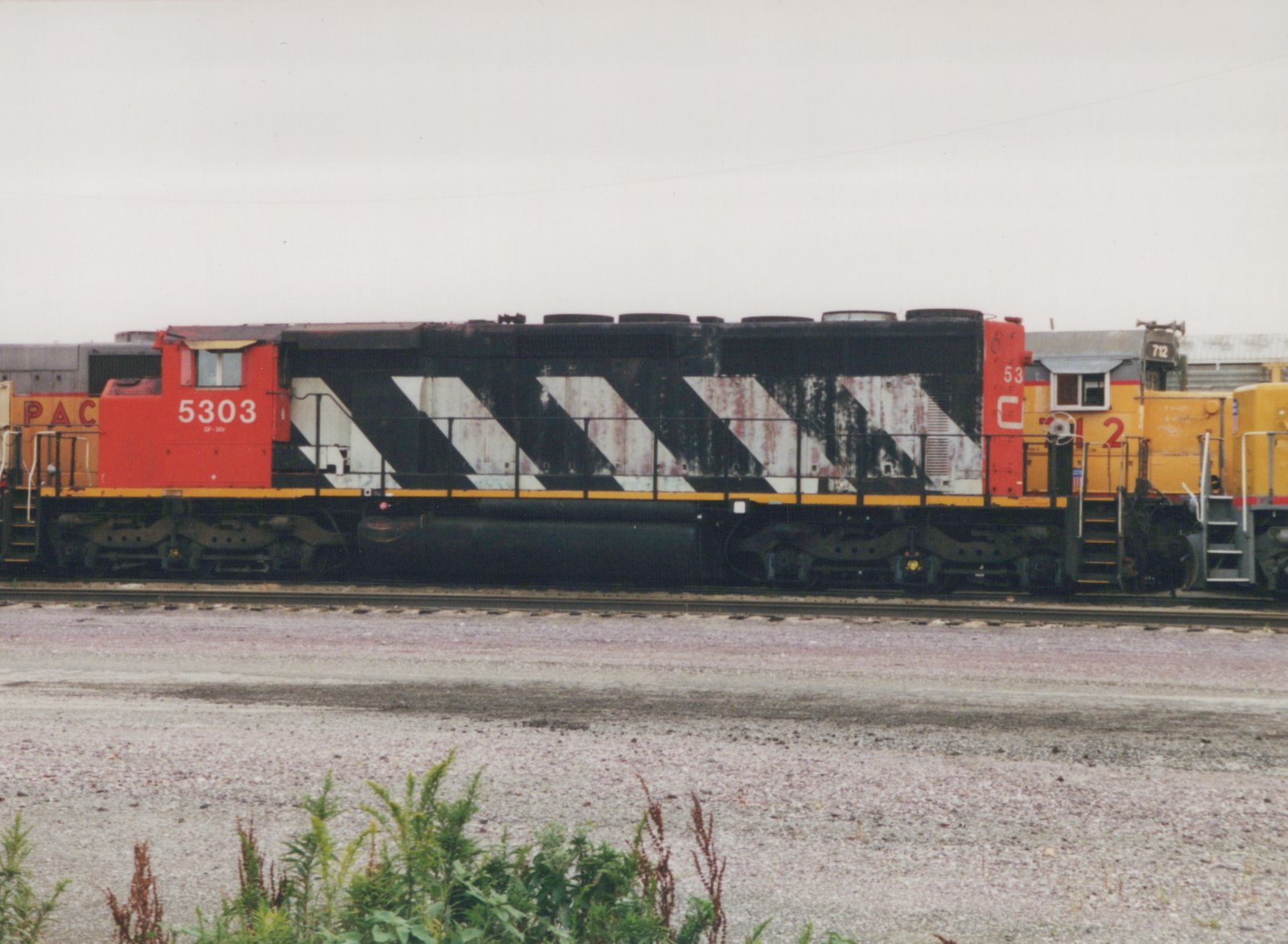 Side view of two diesel locomotives in a gravel yard: a red and black C&NW GP30r with number 5303, and a yellow Pacific Electric SW1200 with number 712, under a gray sky.