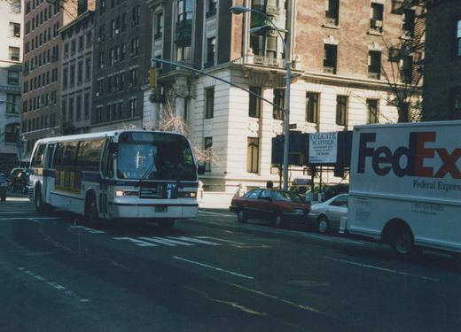 A city bus with the number 4803 and route M5 HOUSTON ST is driving down a street with tall buildings, a FedEx truck, and other cars parked along the side.