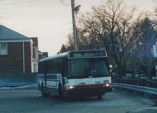 A white, blue and red Chicago Transit Authority bus with the number 5593 is driving on a suburban street with snow on the ground and bare trees in the background during dusk. The destination sign reads '90N to TOUHY'.