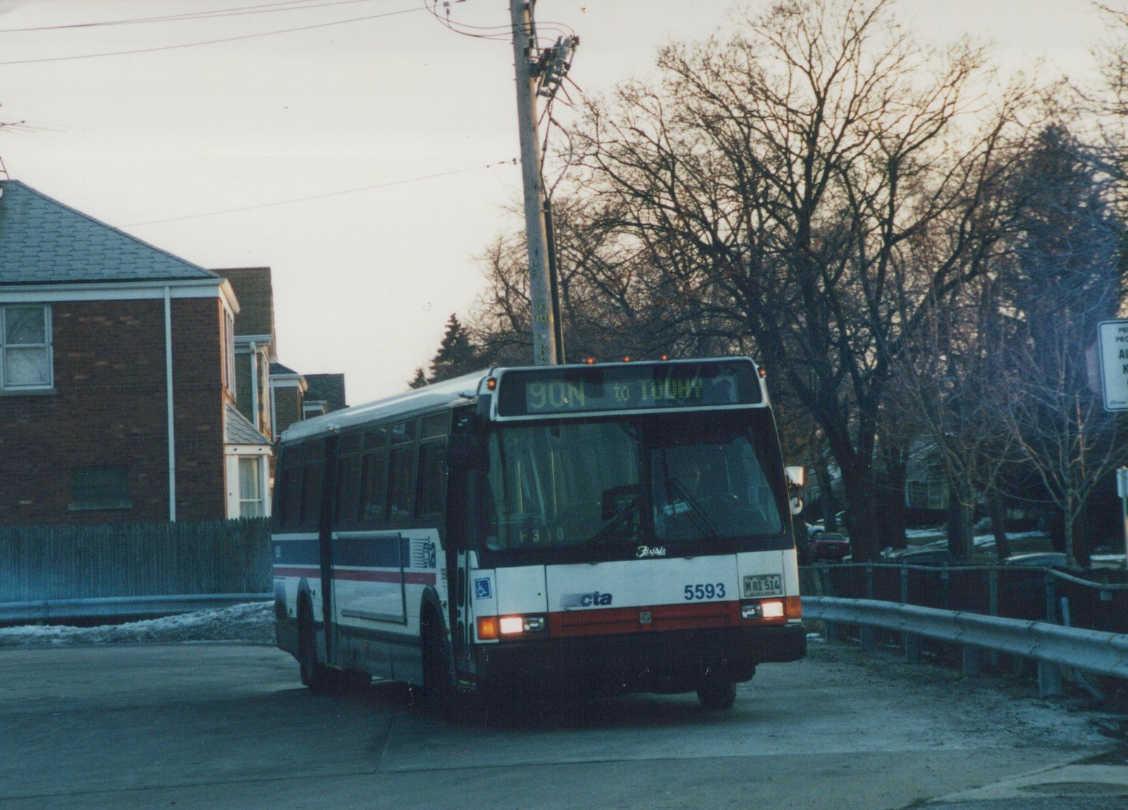 A white, blue and red Chicago Transit Authority bus with the number 5593 is driving on a suburban street with snow on the ground and bare trees in the background during dusk. The destination sign reads '90N to TOUHY'.