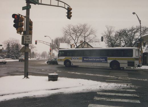 A white Pace bus with a blue and yellow stripe is turning at a snowy street intersection under a traffic light; snow covers the ground and bare trees are in the background.