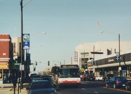 A Chicago Transit Authority bus numbered 5412 drives down a street with a Sears and Homelife Furniture store visible, under a clear sky with an American flag flying.