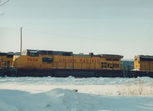 A side view of a yellow Union Pacific locomotive numbered 9668, situated in a snow-covered yard, with other railcars nearby.