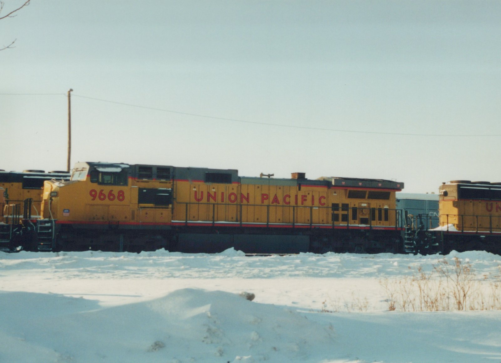 A side view of a yellow Union Pacific locomotive numbered 9668, situated in a snow-covered yard, with other railcars nearby.