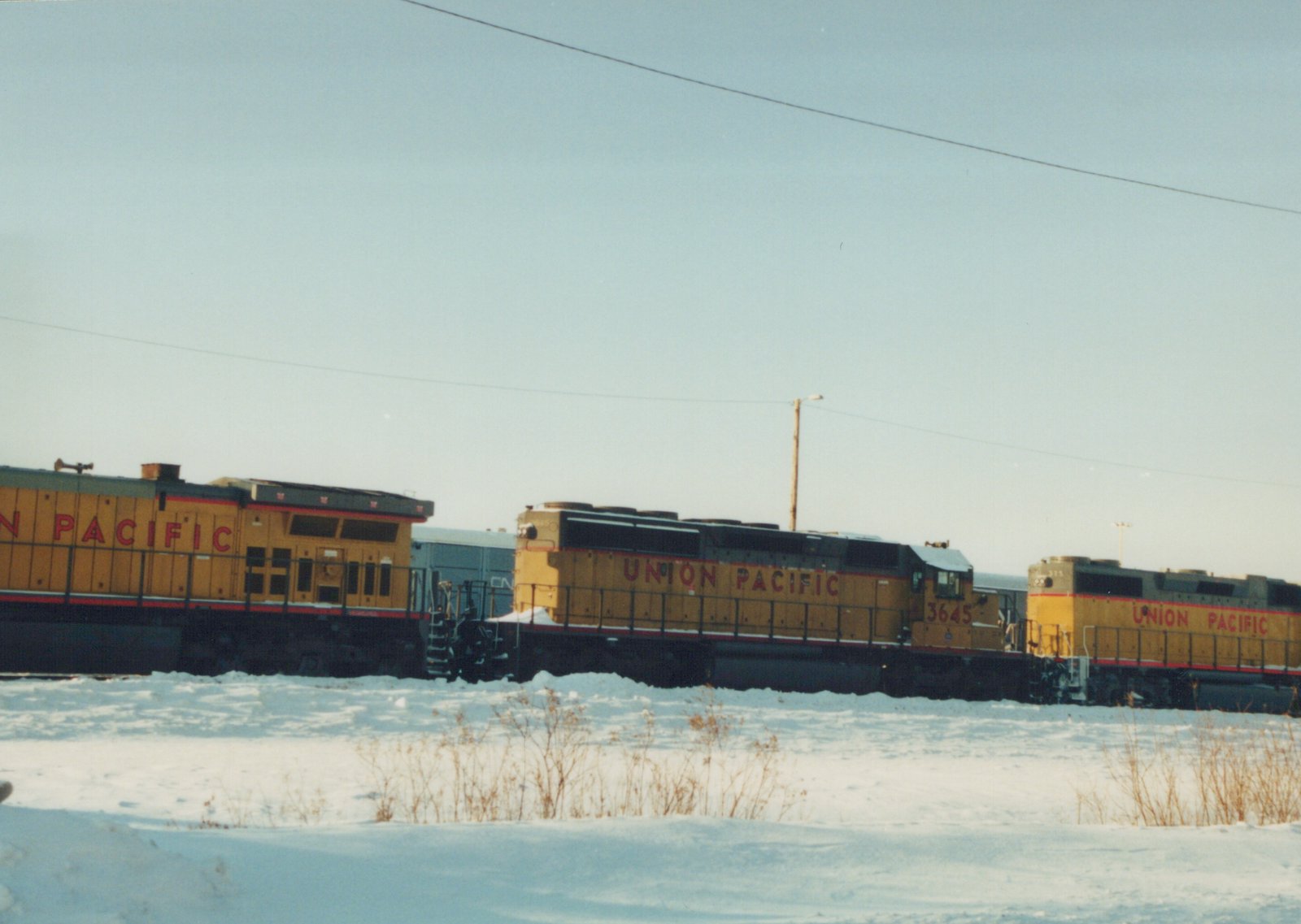 Three yellow and red Union Pacific locomotives, numbers 3645 and 375 visible, are parked side-by-side in a snowy field under a clear sky.