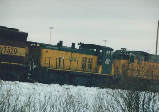 Three yellow and green Chicago and North Western diesel locomotives, numbered 7520, 1307, and 1722, are seen side by side on a snow-covered railroad main line under an overcast sky.