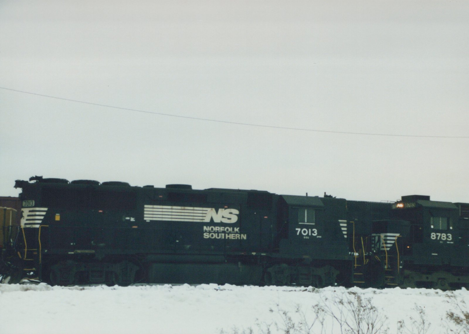 Two black Norfolk Southern diesel locomotives, numbers 7013 and 8783, sit side by side on a snow-covered rail yard under an overcast sky.