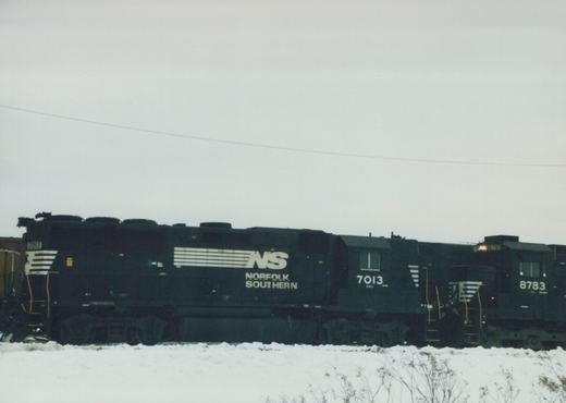 Two black Norfolk Southern diesel locomotives, numbers 7013 and 8783, sit side by side on a snow-covered rail yard under an overcast sky.