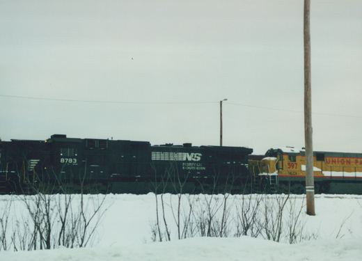 Two diesel locomotives, one black with NS markings and the other yellow with HLCX and Union Pacific lettering, are on a snow-covered track under an overcast sky.
