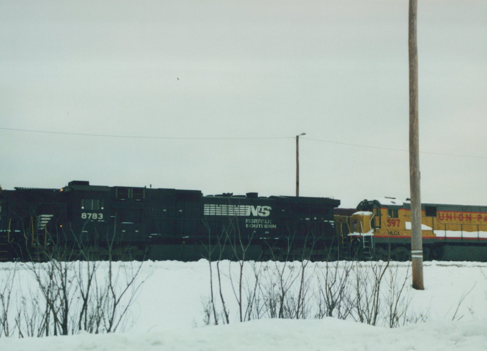 Two diesel locomotives, one black with NS markings and the other yellow with HLCX and Union Pacific lettering, are on a snow-covered track under an overcast sky.