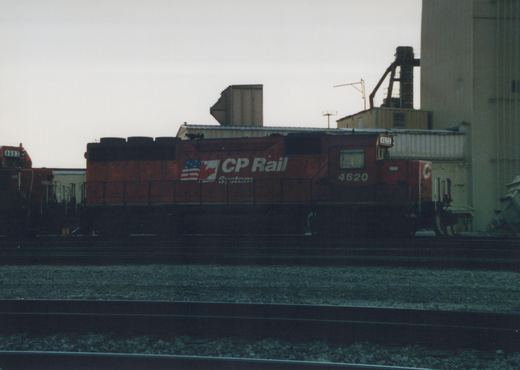 A red CP Rail System diesel locomotive with the number 4620 sits on tracks in a rail yard, with industrial buildings in the background under an overcast sky.