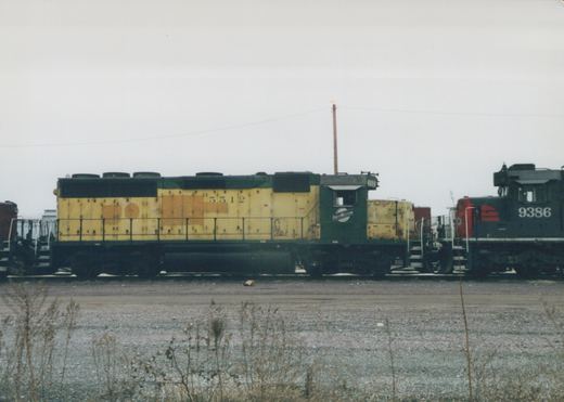 Two diesel locomotives, one yellow and green and the other black and red, are parked side-by-side on railroad tracks in a gravel yard with dry weeds in the foreground under a gray, overcast sky.