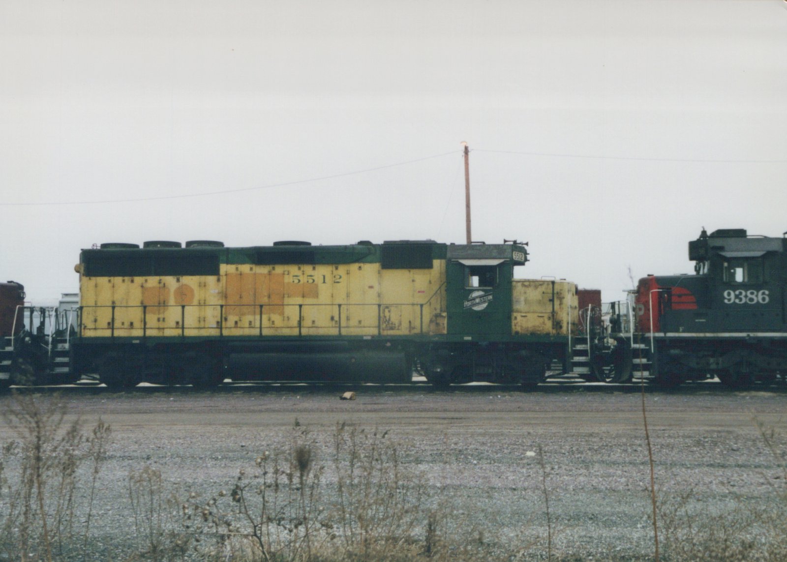 Two diesel locomotives, one yellow and green and the other black and red, are parked side-by-side on railroad tracks in a gravel yard with dry weeds in the foreground under a gray, overcast sky.