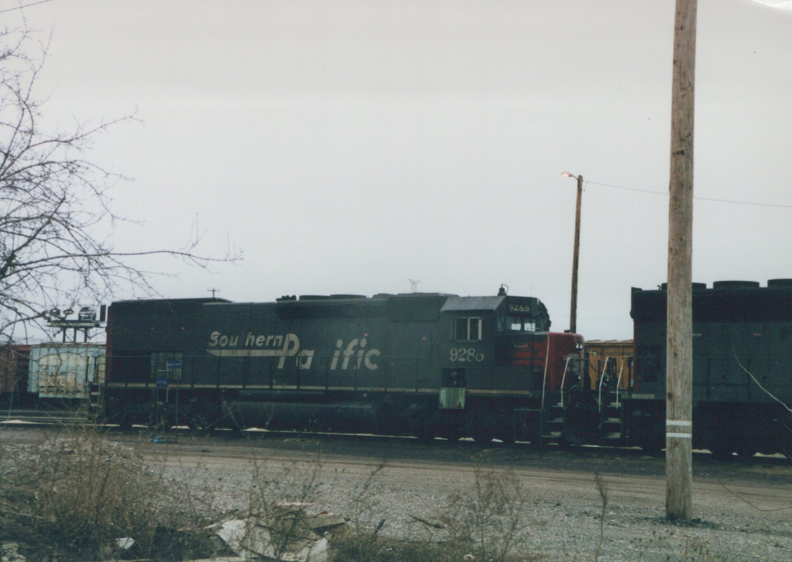 A Southern Pacific diesel locomotive numbered 9288, painted green and red, is parked in a gravel yard with bare trees and utility poles in the background under a gray sky.
