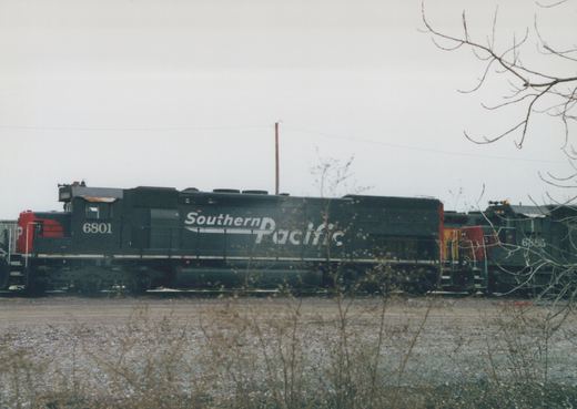 Side view of two Southern Pacific GP38-2 diesel locomotives, numbers 6801 and 6885, parked on a gravel yard with bare trees in the foreground under an overcast sky.