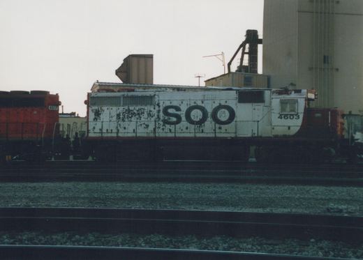 A white and red Soo Line diesel locomotive numbered 4603 sits on tracks in a yard, with industrial buildings in the background.