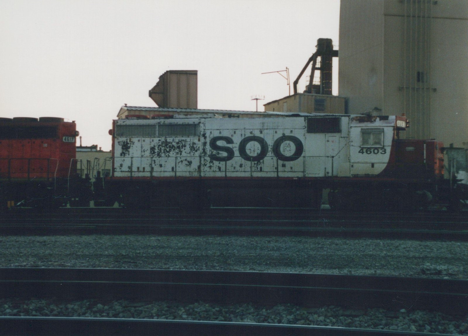 A white and red Soo Line diesel locomotive numbered 4603 sits on tracks in a yard, with industrial buildings in the background.
