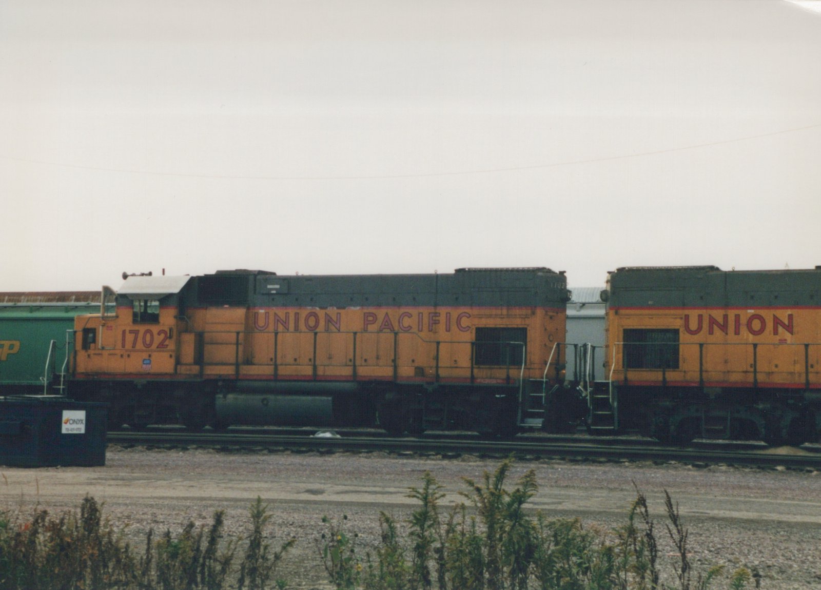 A yellow and black Union Pacific diesel locomotive numbered 1702 is parked on a set of tracks in a yard with overcast skies, with a green freight car partially visible behind it.