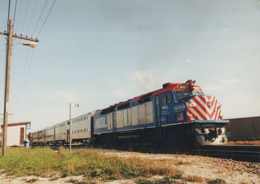 A blue and red Metra locomotive, number 603, pulls a silver double-decker passenger train along tracks next to a grassy area, with a person standing at a small shelter.
