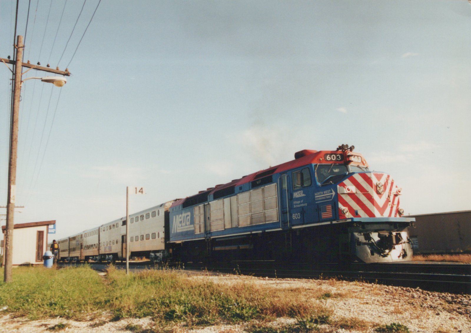 A blue and red Metra locomotive, number 603, pulls a silver double-decker passenger train along tracks next to a grassy area, with a person standing at a small shelter.